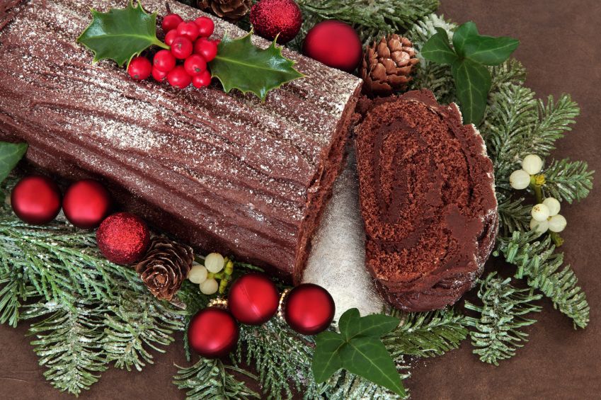 Chocolate yule log cake with red bauble decorations, holly, ivy, mistletoe and snow covered fir over brown handmade lokta paper background.