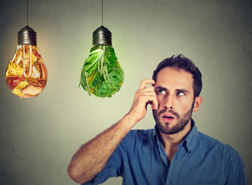 Puzzled man thinking looking up at junk food and green vegetables shaped as light bulbs making decision isolated on gray background. Diet choice right nutrition healthy lifestyle wellness concept