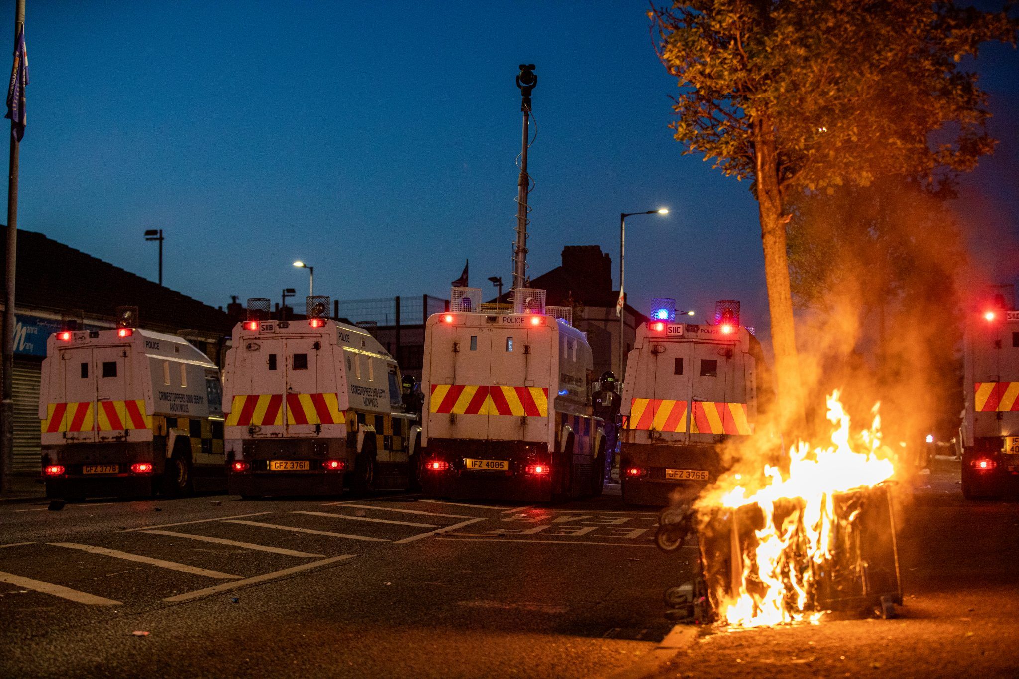 A Biffa bin burns behind a line of riot vans in Belfast